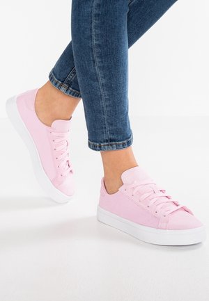 Fødder iført pink snøresneakers kombineret med blå denimjeans afkortet over anklen mod en hvid baggrund.