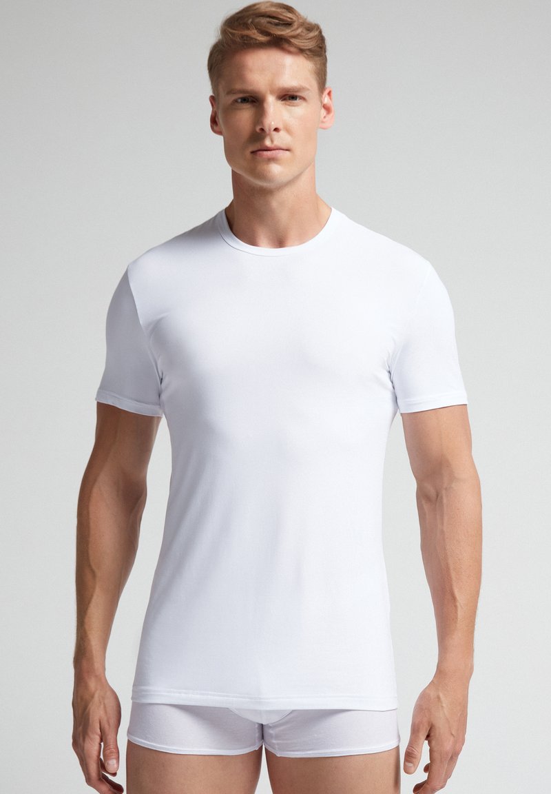 Camiseta blanca de manga corta en una tela suave y ligera con cuello redondo. Llevada con braguitas blancas a juego. Textura suave, diseño ajustado.