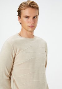 Beige Pullover mit einem strukturierten Wellenmuster, Rundhalsausschnitt und langen Ärmeln, hergestellt aus einem weichen, leichten Material.