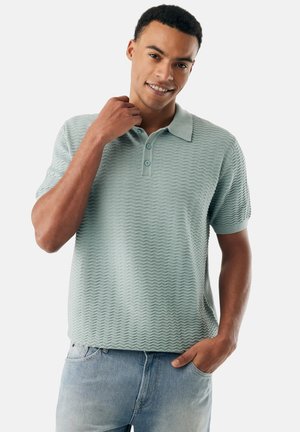 Giovane uomo sorridente, indossa una polo a maniche corte verde chiaro con texture e jeans azzurri, con una mano in tasca e l'altra vicino al colletto.