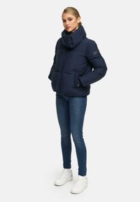 Vrouw die met handen in de zakken staat, gekleed in een marineblauwe pufferjack, blauwe skinny jeans en witte sneakers op een effen achtergrond.