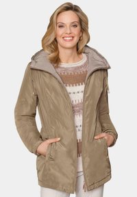 Veste imperméable beige avec une grande capuche, fermeture éclair et poches latérales ; possède une texture lisse et des poignets élastiques. Portée par-dessus un pull à motifs.