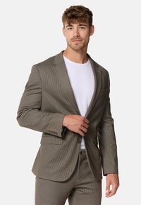 Blazer a quadri marrone con una texture liscia, chiusura a un bottone e rever a scollo a V, indossato sopra una maglietta bianca.