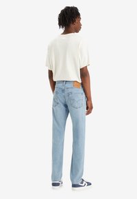 Uomo con dreadlocks che indossa una maglietta bianca, jeans azzurri chiari e sneaker blu navy, in piedi di spalle su uno sfondo semplice.