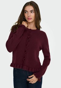 Pull bordeaux avec un col rond, boutonnière à l'avant et des volants au bas et aux poignets. Fabriqué en tissu tricoté doux et texturé.