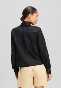 Schwarzes Langarmshirt mit hohem Kragen, glatter Textur und geknöpften Manschetten; verfügt über eine Mittelnaht hinten und eine taillierte Passform.