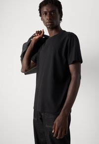 Man in een zwart T-shirt en donkere spijkerbroek draagt een zwarte tas over zijn schouder tegen een effen lichte achtergrond.