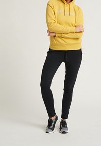 Geel hoodie met witte logo, gecombineerd met zwarte slim-fit joggers met zijzakken en zwarte sneakers met grijze accenten.