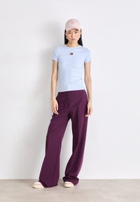 Maglietta aderente azzurro chiaro con un piccolo logo, abbinata a pantaloni larghi viola scuro e scarpe con zeppa beige; un berretto rosa completa il look.