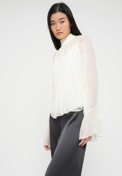 Blouse blanche plissée avec un col montant et des manches évasées. Tissu léger et transparent avec des cordons de serrage aux extrémités des manches. Associée à un pantalon foncé.