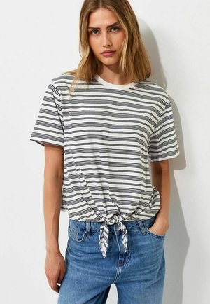 Jonge vrouw draagt een wit T-shirt met korte mouwen met zwarte horizontale strepen, voorgebonden, en een blauwe spijkerbroek met één hand in de zak.