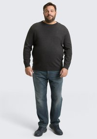 Donkergrijze trui met een ronde hals, gedragen over lichtblauwe jeans en zwarte schoenen. De stof lijkt glad met een subtiele textuur.
