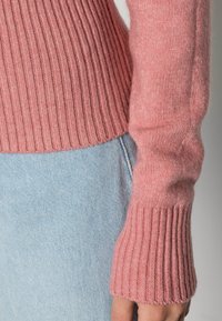 Nära bild av en person som bär en ribbad rosa tröja med ärmar i kombination med ljusblå denimjeans.