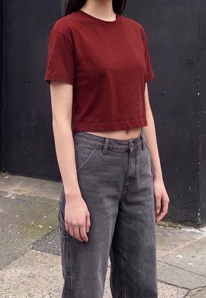 Personne portant une chemise courte rouge-brun à manches courtes et un jean gris taille haute, debout dehors contre un mur sombre.