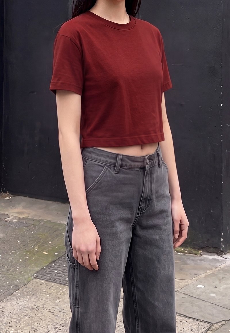 Personne portant une chemise courte rouge-brun à manches courtes et un jean gris taille haute, debout dehors contre un mur sombre.