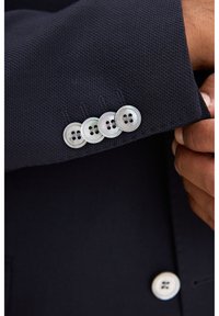 Mörkblå texturerad kavajärmsmanschett med fyra vita knappar med två hål vardera, hålls av en hand som justerar tyget.