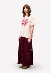 T-shirt en coton beige avec un motif graphique rouge, assorti à une jupe longue en dentelle bordeaux ornée de motifs de feuilles. Le modèle porte des chaussons marron.