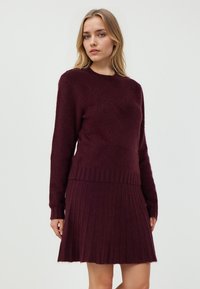 Burgunderfarbendes Strickkleid mit langen Ärmeln, geripptem Saum und strukturiertem Oberteil, das einen plissierten Rock hat. Weicher, warmer Stoff.