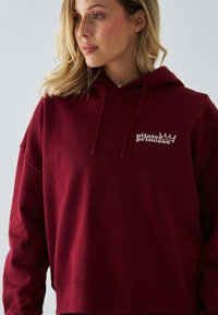 Burgundowa bluza z kapturem o luźnym kroju, z napisem "królowa pilates" w białym kolorze oraz małą grafiką korony. Miękka tkanina i minimalistyczny design.