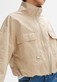 Beige verkürzte Jacke aus leichtem Stoff, mit zwei vorderen Taschen, hohem Kragen und einem silbernen Reißverschluss. Strukturiert Oberfläche hebt die Details hervor.