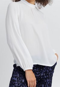 Blusa bianca a maniche lunghe in tessuto leggero con collo alto, dettagli plissettati e polsini elastici. Abbinata a pantaloni blu scuro con fantasia.