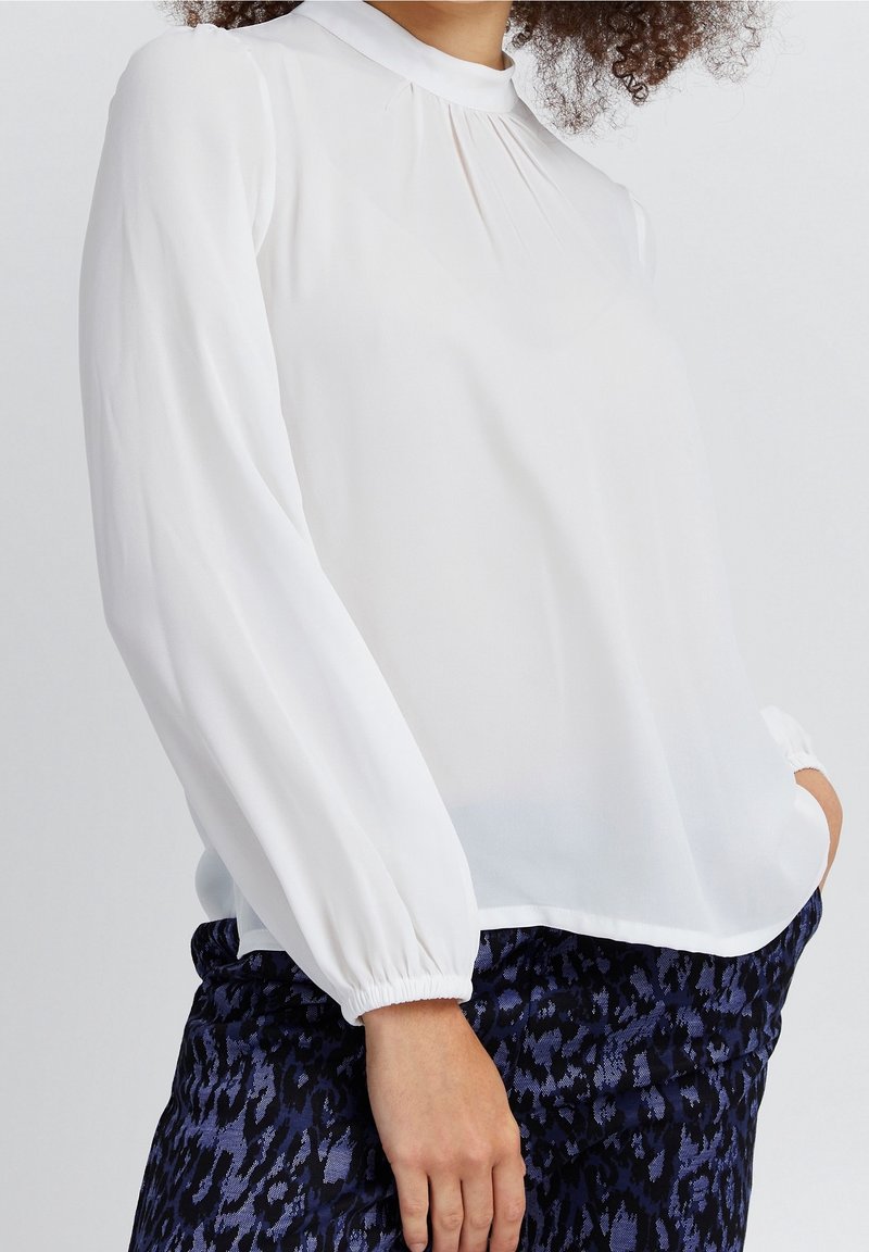 Blusa bianca a maniche lunghe in tessuto leggero con collo alto, dettagli plissettati e polsini elastici. Abbinata a pantaloni blu scuro con fantasia.