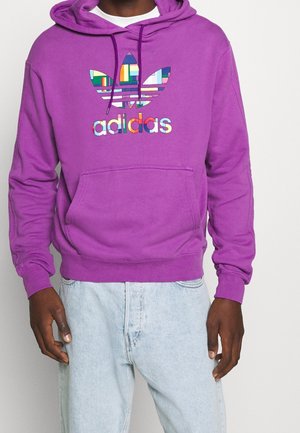 Haut à capuche violet avec une poche avant, arborant un logo trefoil coloré d'Adidas. Le tissu semble doux avec une finition texturée.