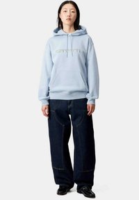 Sudadera azul claro con un bolsillo frontal tipo canguro y capucha con cordones, combinada con pantalones de mezclilla oscuros de gran tamaño que presentan costuras amarillas.