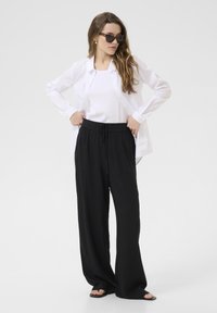 Camicia bianca a bottoni, canotta bianca aderente, pantaloni neri a gamba larga e sandali neri. La modella indossa occhiali da sole rettangolari; capelli lunghi, senza motivi visibili.