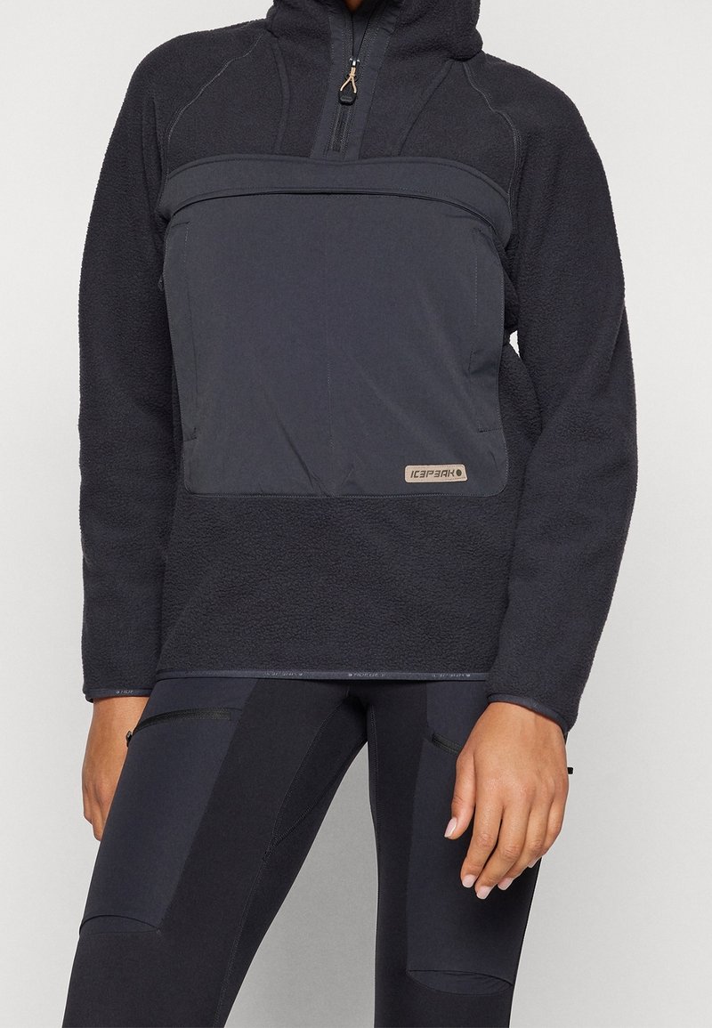 Sweat à capuche en polaire noire avec poche zippée à l'avant, porté avec des leggings noirs avec poches latérales zippées.