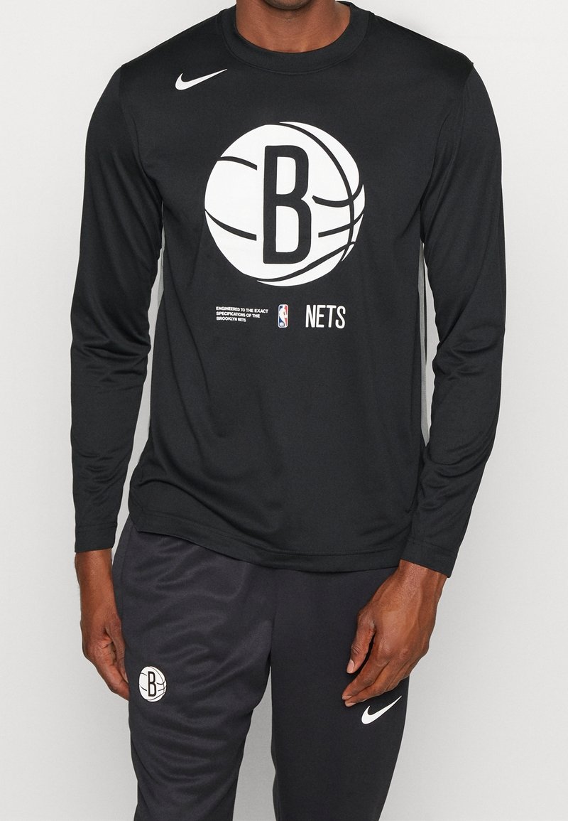 Schwarzes, langärmliges Nike-Shirt mit einem großen weißen Brooklyn Nets-Logo und Text darunter; kombiniert mit schwarzen Hosen, die ein Logo zeigen.
