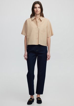 Light beige cropped button-up shirt with short sleeves, featuring a pocket with perforated detailing. Paired with dark blue straight-leg jeans.