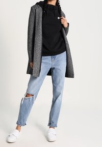 Grå lång kappa över en svart hoodie, tillsammans med ljusblå slitna jeans och vita slip-on skor. Enkel och avslappnad outfitkomposition.