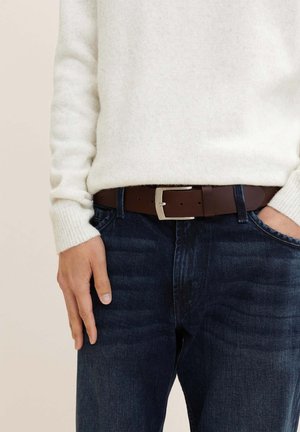 Ceinture en cuir marron foncé avec une boucle en métal argenté, portée sur un jean en denim foncé, avec une manche de pull crème clair visible.