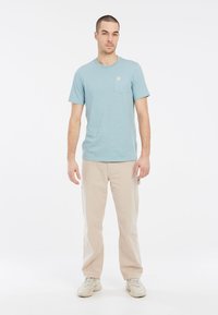 Svetlomodré tričko s krátkymi rukávmi a vreckom na hrudi v kombinácii s béžovými nohavicami. Tkanina vyzerá jemne a mierne štruktúrovaná.