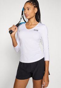 Top atlético branco de manga comprida com decote em V e logótipo preto, combinado com calções pretos. A segur uma raquete de ténis com um detalhe azul.