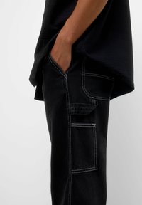 Persona vistiendo pantalones negros con costuras blancas, con una mano en el bolsillo delantero y una camiseta negra texturizada, mostrada de lado sobre un fondo blanco.