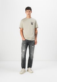 Béžové bavlněné tričko s černým grafickým logem, spárované s opranými šedými džíny. Bílé tenisky dotvářejí celý outfit.