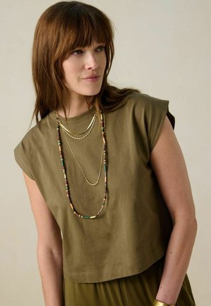 Olijfgroene cropped t-shirt met opgerolde korte mouwen, gecombineerd met gelaagde kettingen met meerkleurige kralen, textuurstof en een ontspannen pasvorm.