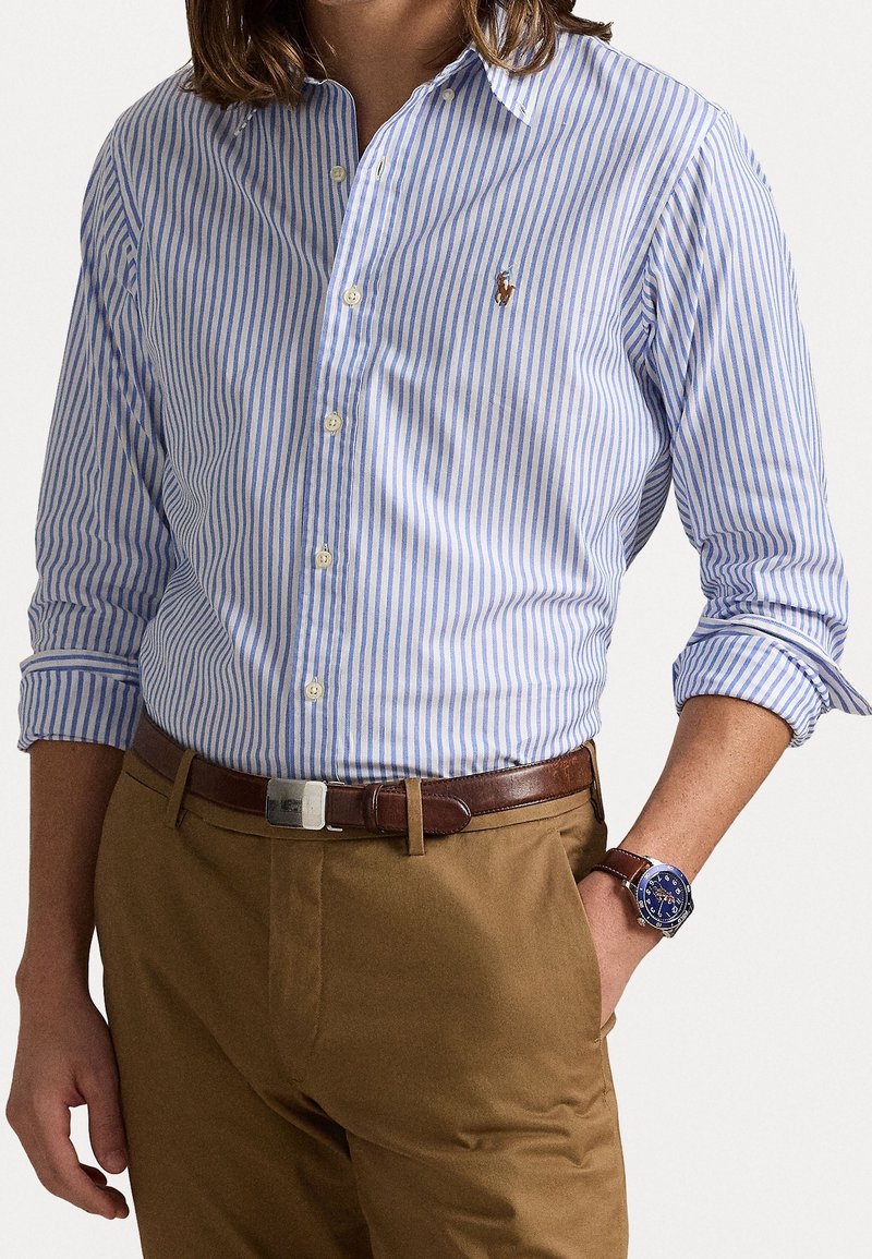 Blau-weiß gestreiftes Hemd mit langen Ärmeln, kombiniert mit braunen Hosen und einem braunen Leder-Gürtel sowie einer Uhr.