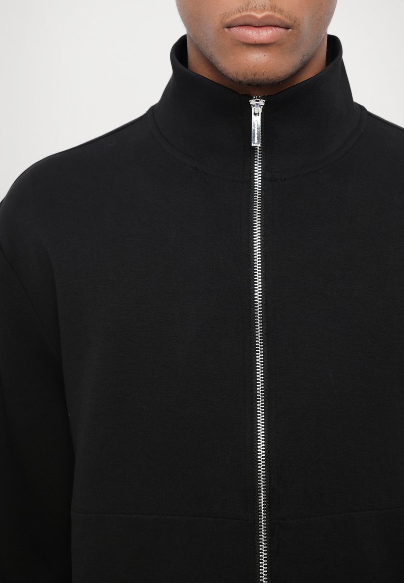 Giacca nera con zip realizzata in tessuto misto cotone, caratterizzata da un colletto alto e da una zip in tonalità argento davanti, con una texture liscia.
