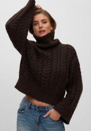 Femme portant un pull à col roulé torsadé marron foncé et un jean bleu, une main dans la poche et l’autre levée au-dessus de sa tête.