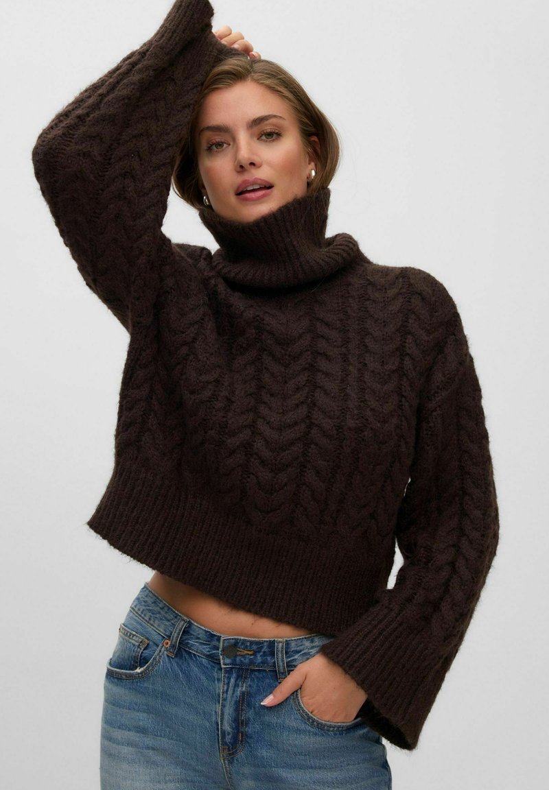 Femme portant un pull à col roulé torsadé marron foncé et un jean bleu, une main dans la poche et l’autre levée au-dessus de sa tête.