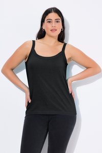 Woman with long dark hair wearing black sleeveless top and dark pants, standing with hands on hips against white background.