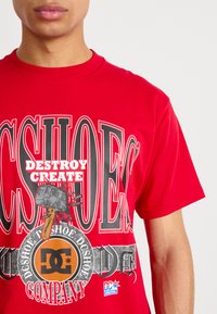 T-shirt graphique rouge avec du texte noir et blanc en gras, présentant un logo central, un graphique de marteau et une petite étiquette de marque sur le côté inférieur.