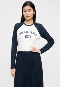 Camisa de manga comprida com corpo branco e mangas raglan azul-marinho com estampa "MICHAEL KORS 1981". Combinada com saia plissada azul-marinho.