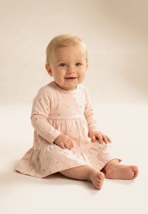Smiješeća beba sa svijetlom kosom sjedi, noseći blijedo rozu haljinu s dugim rukavima i malim uzorcima srca na neutralnoj pozadini.