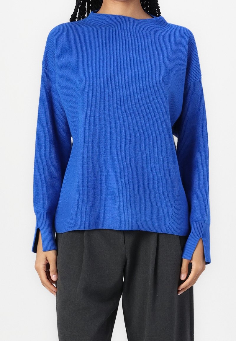 Personne portant un pull en maille côtelée bleu vif avec des poignets fendus et un pantalon ample gris foncé sur un fond blanc uni.