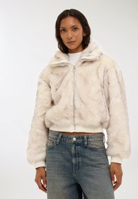 Giacca in pelliccia sintetica beige chiaro con zip frontale, colletto alto e vita a coste. Indossata sopra jeans in denim sbiaditi a vita alta.