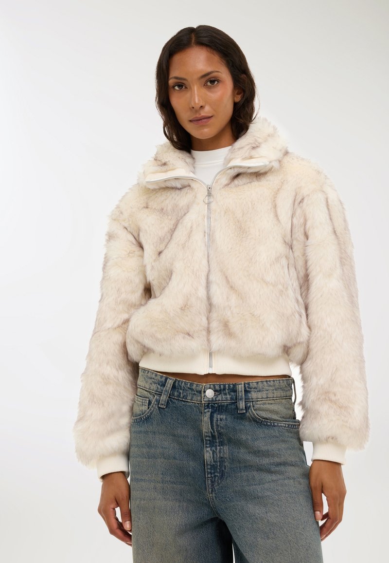 Giacca in pelliccia sintetica beige chiaro con zip frontale, colletto alto e vita a coste. Indossata sopra jeans in denim sbiaditi a vita alta.